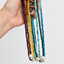 A hand with dark nail polish holds up a bundle of long beaded necklaces in multiple colors, including deep red, yellow, green, teal, and blue, with white pearl-like beads at the ends, against a plain light gray background.
