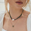A close-up of a woman wearing layered necklaces, including a dark green beaded choker with a white pearl, a gold sunburst necklace with a green center, and a delicate gold chain.