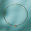 A delicate gold wire hoop necklace with a small heart-shaped pendant, displayed against a soft teal background with leaf shadows.